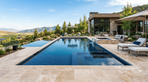 A sprawling modern-mountain home features a luxury pool deck meticulously crafted from light, oversized travertine tiles. This elegant stone surface surrounds a geometric infinity pool and attached spa, providing a cool and stable surface that complements the panoramic Utah mountain valley views.
