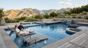 A couple relaxes on submerged chaise lounges within a modern luxury pool, which features a tanning ledge, built-in social benches, and a spa overlooking mountains. The scene showcases integrated seating options that transform the high-end aquatic space into a comfortable social retreat.